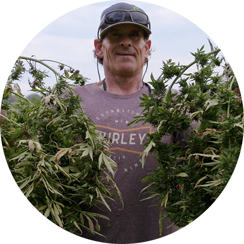 Bo Halley standing outside smiling and holding a large bush of hemp in each hand.