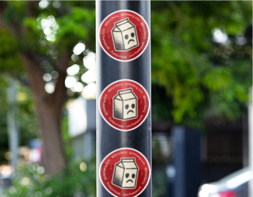Photograph of a pole with two round stickers bearing an image of a milk carton with a sad face encircled by a campaign hashtag and URL.
